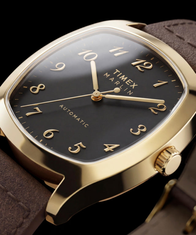 Közeli kép az aranyszínű Timex Marlin Chatham Automatic óráról, amelyen látható a fekete számlap, a rátett aranyszínű arab számok, az aranyszínű mutatók és a barna bőrszíj