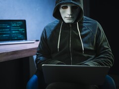 Egy maszkos, csuklyás alak dolgozik egy laptopon egy sötét szobában.