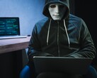 Egy maszkos, csuklyás alak dolgozik egy laptopon egy sötét szobában.