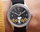 A Timex MK1 fekete számlapjának közeli képe, amelyen Snoopy látható egy sárga-fekete kockás mintán, arab számokkal jelölt órajelölőkkel és világító mutatókkal.
