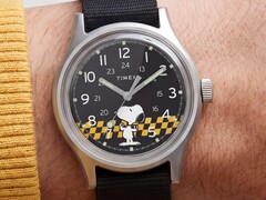 A Timex MK1 fekete számlapjának közeli képe, amelyen Snoopy látható egy sárga-fekete kockás mintán, arab számokkal jelölt órajelölőkkel és világító mutatókkal.