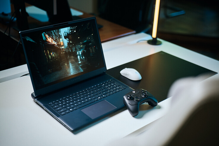 Egy kinyitott Alienware gamer laptop egy fehér stúdióasztalon, amelyen egy sötét, tükröződő utcai jelenet fut éles, path-traced részletességgel, mellette egy fekete vezérlő és egy fehér vezeték nélküli egér.