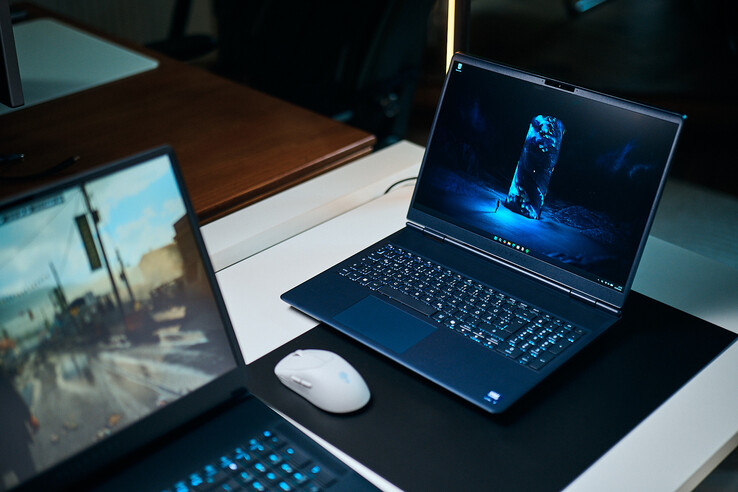 Egy sötétkék, középkategóriás gamer laptop (Alienware Aurora 16X) ferde szögben áll egy íróasztali alátéten, amelyen ragyogó kék, sci-fi ihletésű háttérkép látható; mellette egy fehér vezeték nélküli egér fekszik, az előtérben pedig egy másik laptop látható elmosódva.