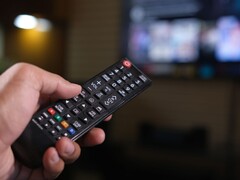Távirányítót tartó kéz, a háttérben elmosódott tévével
