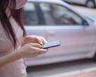 Egy nő az okostelefonján gépel, a háttérben egy ezüstszínű autóval