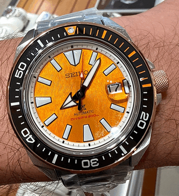 A Seiko Prospex HBB005K1 közeli felvétele, amelyen látható a texturált borostyánszínű számlap, a túlméretezett, világító órajelölők, a széles mutatók, a dátumablak és a narancssárga jelölésekkel ellátott fekete búvárkeret.