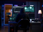 Egy csuklyás kutató több képernyőn dolgozik egy sötétített berendezésben. Általános hacker a képen. 