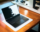 Egy nyitott, matt fekete Razer Blade 16 gamer laptop, amely egy fehér asztali szőnyegen fekszik egy meleg hangulatú faasztalon, élénk fekete-fehér háttérképpel.