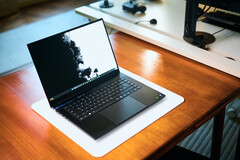 Egy nyitott, matt fekete Razer Blade 16 gamer laptop, amely egy fehér asztali szőnyegen fekszik egy meleg hangulatú faasztalon, élénk fekete-fehér háttérképpel.