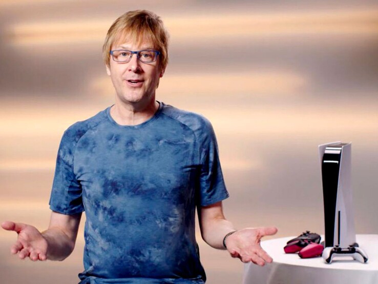Mark Cerny egy PlayStation eseményen, egy PS5 konzol és DualSense kontrollerek mellett ülve. (Kép forrása: Sony Interactive Entertainment)
