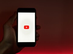 Egy kéz, amelyben egy okostelefon van, amelynek kijelzőjén a YouTube logó világít vörös háttér előtt
