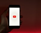 Egy kéz, amelyben egy okostelefon van, amelynek kijelzőjén a YouTube logó világít vörös háttér előtt