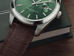 Közeli kép a Timex Legacy Day and Date óráról, amelyen látható a zöld, napsugármintás számlap, az ezüstszínű lünetta és a szálcsiszolt tokfülek, a bordázott korona, valamint a texturált barna bőrszíj, sötétzöld dobozzal és fafelülettel a háttérben.