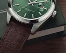 Közeli kép a Timex Legacy Day and Date óráról, amelyen látható a zöld, napsugármintás számlap, az ezüstszínű lünetta és a szálcsiszolt tokfülek, a bordázott korona, valamint a texturált barna bőrszíj, sötétzöld dobozzal és fafelülettel a háttérben.