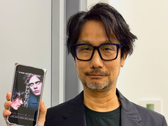 Hideo Kojima a Physint poszterét mutatja a telefonján