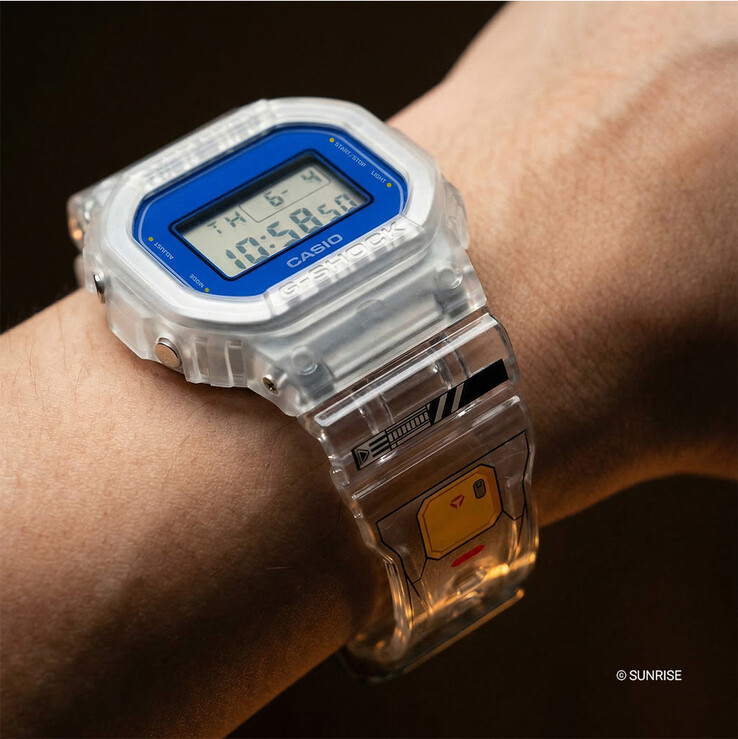 Casio G-Shock x Mobile Suit Gundam DW-5600GM26-7DR csuklón viselve, átlátszó műgyanta tokkal és szíjjal, kék lünetta körítéssel és Gundam ihlette grafikával a szíjon.