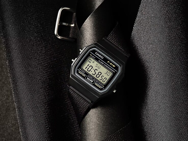 Casio F-91WF-1A digitális karóra fekete tokban, fekete szövött szíjjal és ezüst díszítéssel, sötét, texturált háttér előtt fényképezve.