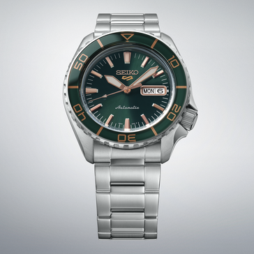Seiko 5 Sports SKX HDB005 sötétzöld számlappal, hozzá illő zöld lünettával, rózsaarany színű mutatókkal és indexekkel, valamint rozsdamentes acél szíjjal, világosszürke háttér előtt.