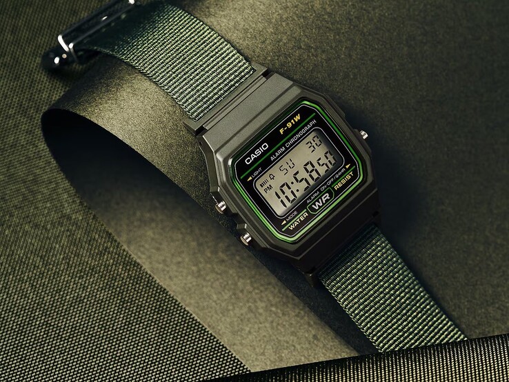 Casio F-91WF-3A digitális karóra, zöld díszítésű fekete tokban és zöld szövött szíjjal, sötétzöld, texturált háttér előtt fényképezve.