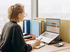 Szemüveges nő laptopon táblázatkezelő programmal dolgozik, az asztalon iratgyűjtők és egy számológép található