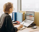 Szemüveges nő laptopon táblázatkezelő programmal dolgozik, az asztalon iratgyűjtők és egy számológép található
