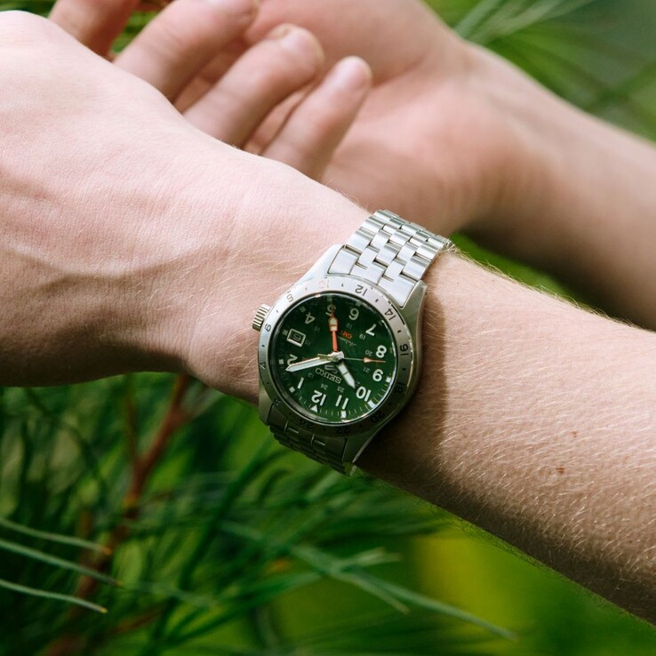 A Seiko 5 Sports Field GMT HDB001 modell elölnézete: zöld számlappal, rögzített rozsdamentes acél 24 órás kerettel, narancssárga GMT-mutatóval és ötszemű acél karkötővel.
