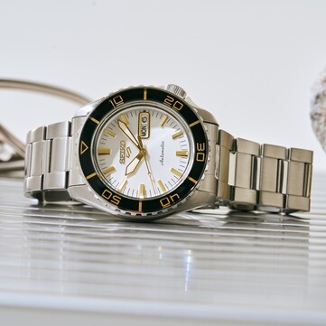 Seiko 5 Sports SKX HDB004 karóra ezüstfehér számlappal, fekete forgatható lünettával és aranyszínű jelölésekkel, aranyszínű mutatókkal és indexekkel, valamint rozsdamentes acél szíjjal, ferdén fényképezve.