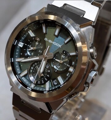 Közelkép a zöld számlapos Casio G-Shock GST-W1000D-3A óráról, amelyen látható a geometrikus textúrájú számlap, a három kis számlap, a világító jelölések és a csiszolt rozsdamentes acél keret.