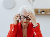 Egy idősebb, fehér hajú, szemüveges nő telefonál egy világos nappaliban, miközben kezét a halántékához szorítja.