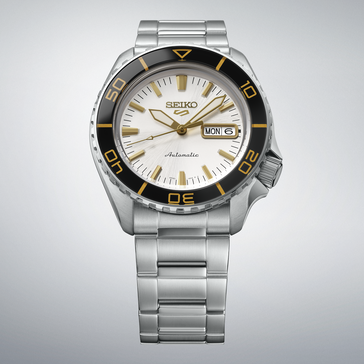 Seiko 5 Sports SKX HDB004 ezüstfehér számlappal, fekete lünettával és aranyszínű jelölésekkel, aranyszínű mutatókkal és indexekkel, valamint rozsdamentes acél szíjjal, világosszürke háttér előtt.